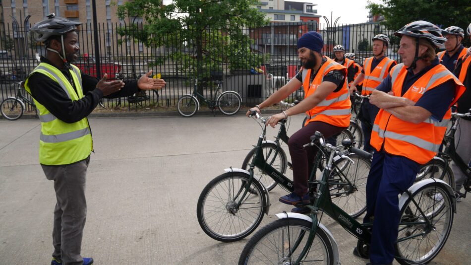 Group of lorry drivers in orange hi-vis and on cycles receiving instruction from a cycle instructor