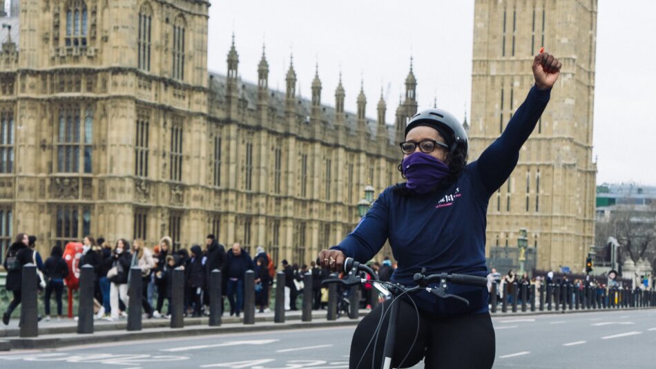 Woman of colour cycling past Parliament wearing helmet and navy top and black trousers, raising left hand in a fist