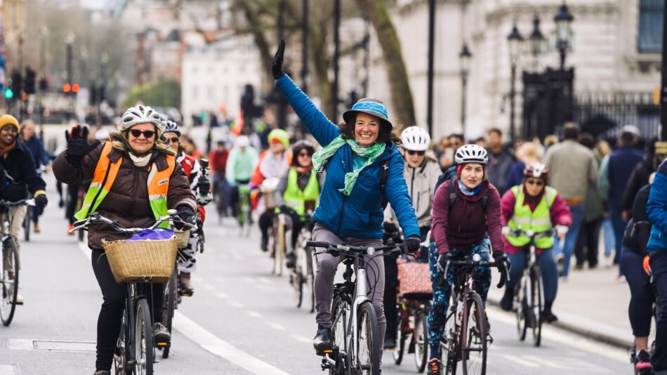 A group of women on bicycles ride towards the camera - in the centre one woman is raising her arm above her head and smiling